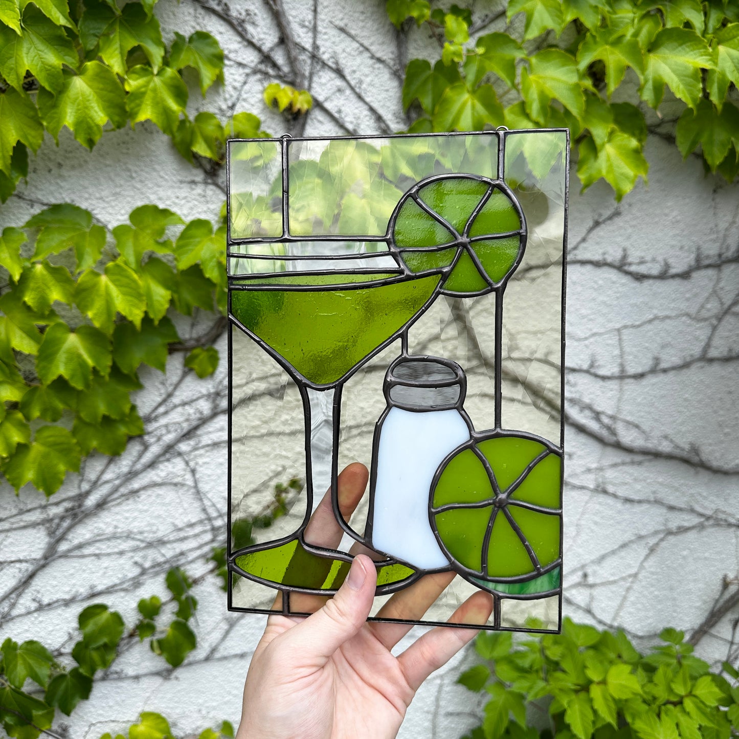 Stained glass panel of a cocktail with lime and salt shaker held against a leafy background
