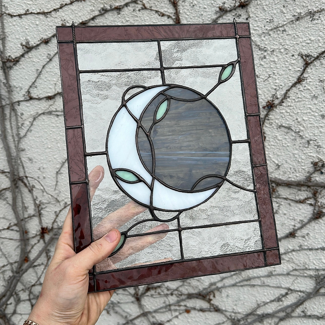 A stained glass window of a white moon wrapped in green vines with a purple border.