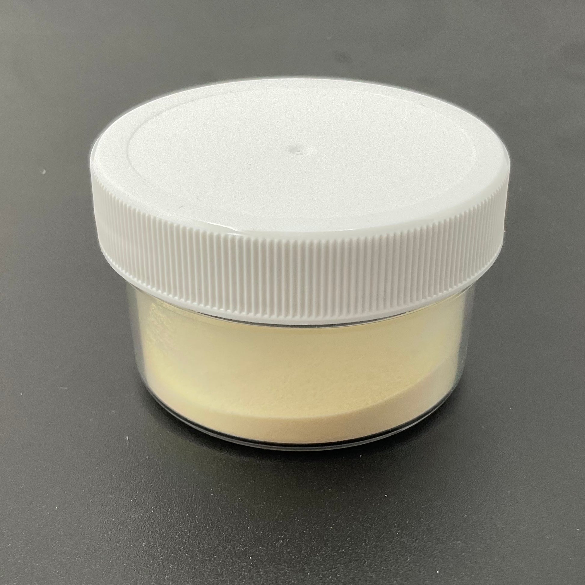 Small clear container with beige powder and a white lid on a dark gray background