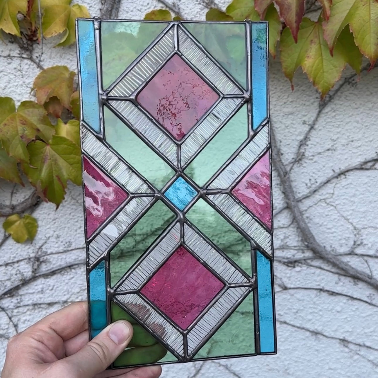 Hand holding a geometric stained glass panel with pink, blue, and green colors against a textured wall and leafy background.