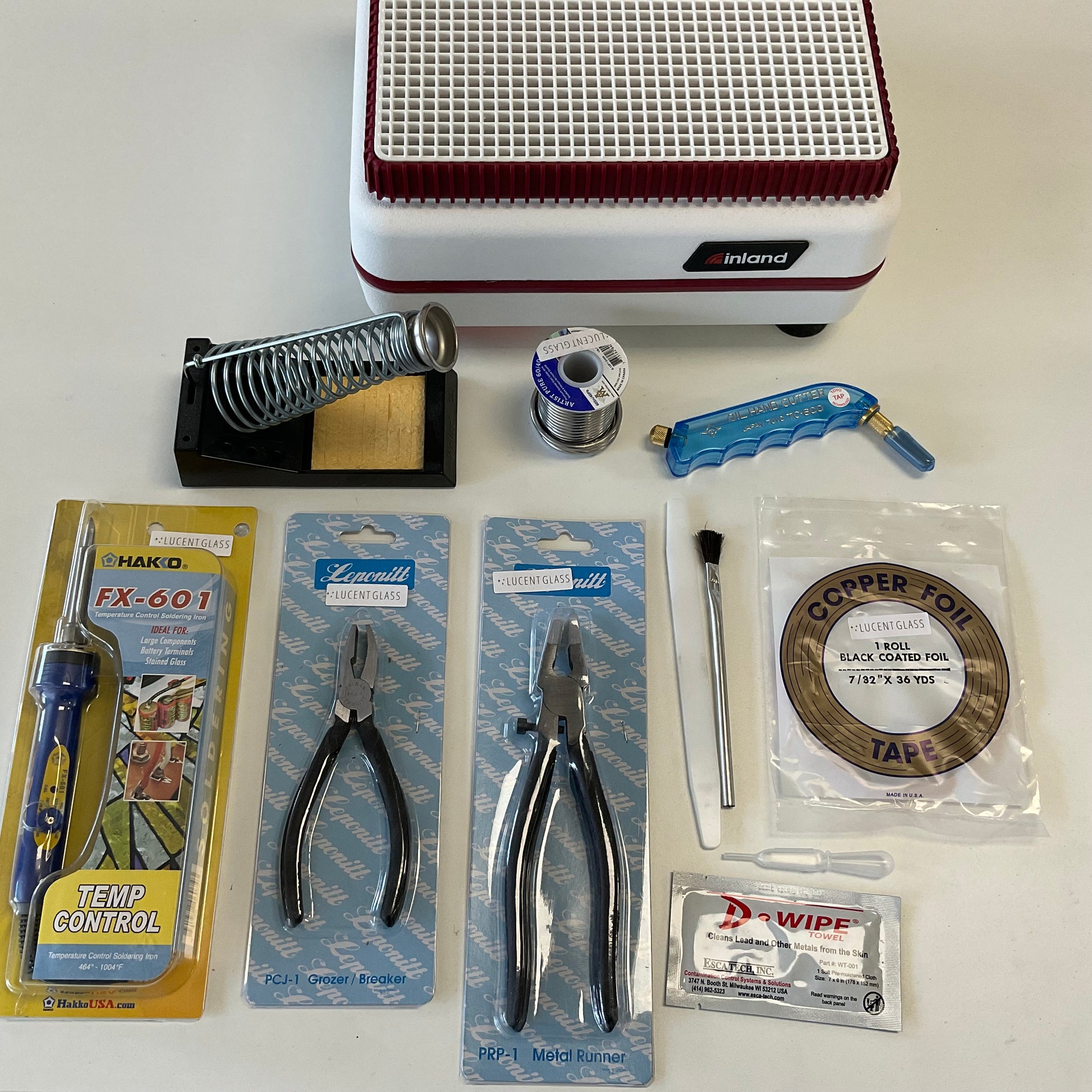 A collection of stained glass making tools and materials laid out on a table, including grinders, pliers, a glass cutter, soldering irons, foil, flux, patina, and a measuring square.